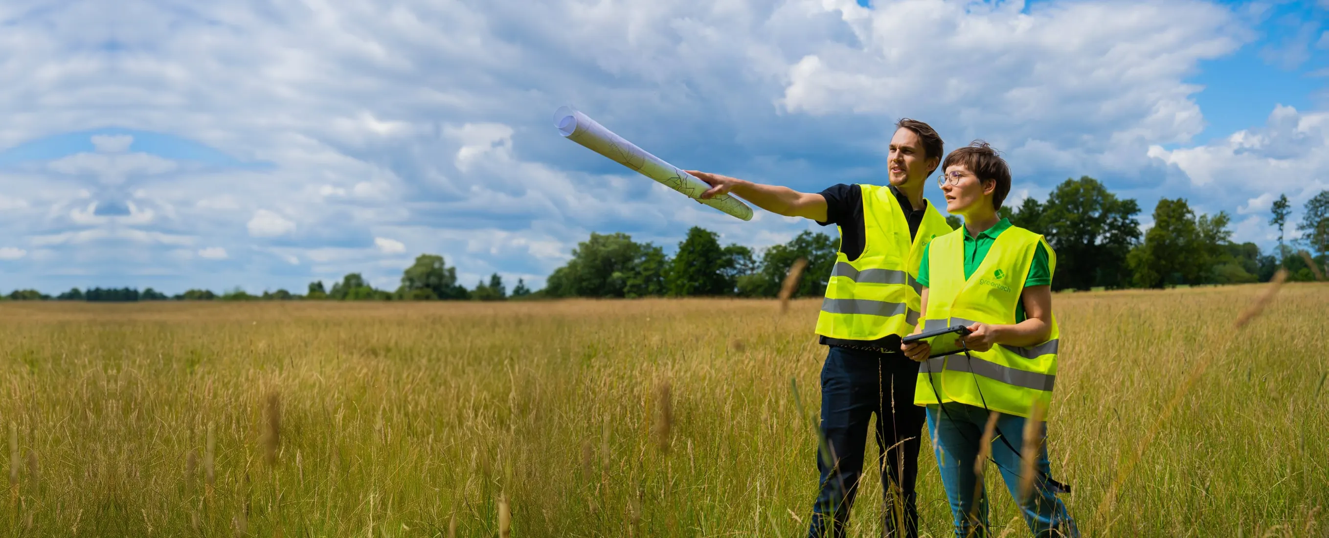 Two greentech project developers in high-visibility vests stand in a summer meadow. One points across the land with a rolled-up site plan, the other holds a tablet.