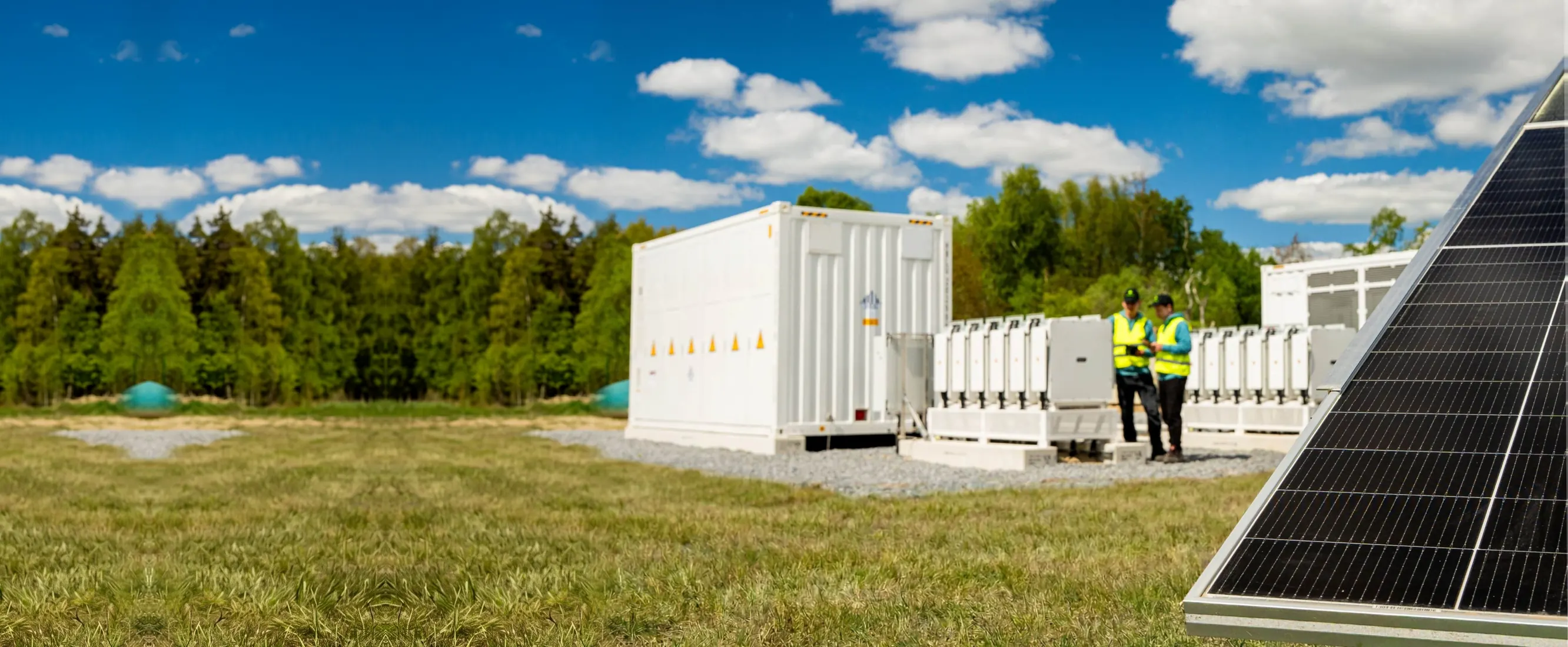 White battery storage containers and inverters on gravel, two greentech engineers in high-visibility vests between them, the edge of a solar module in the foreground.