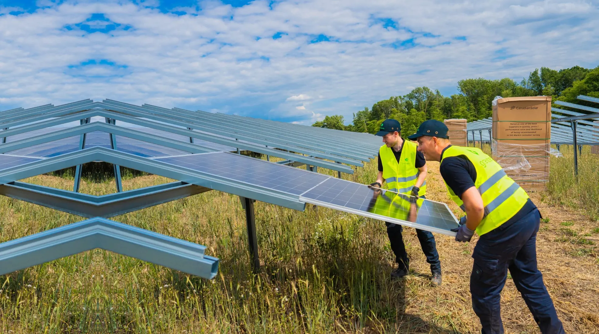 Two greentech installers in high-visibility vests lift a solar module onto a steel mounting frame. Pallets of modules stand behind them under a blue sky.