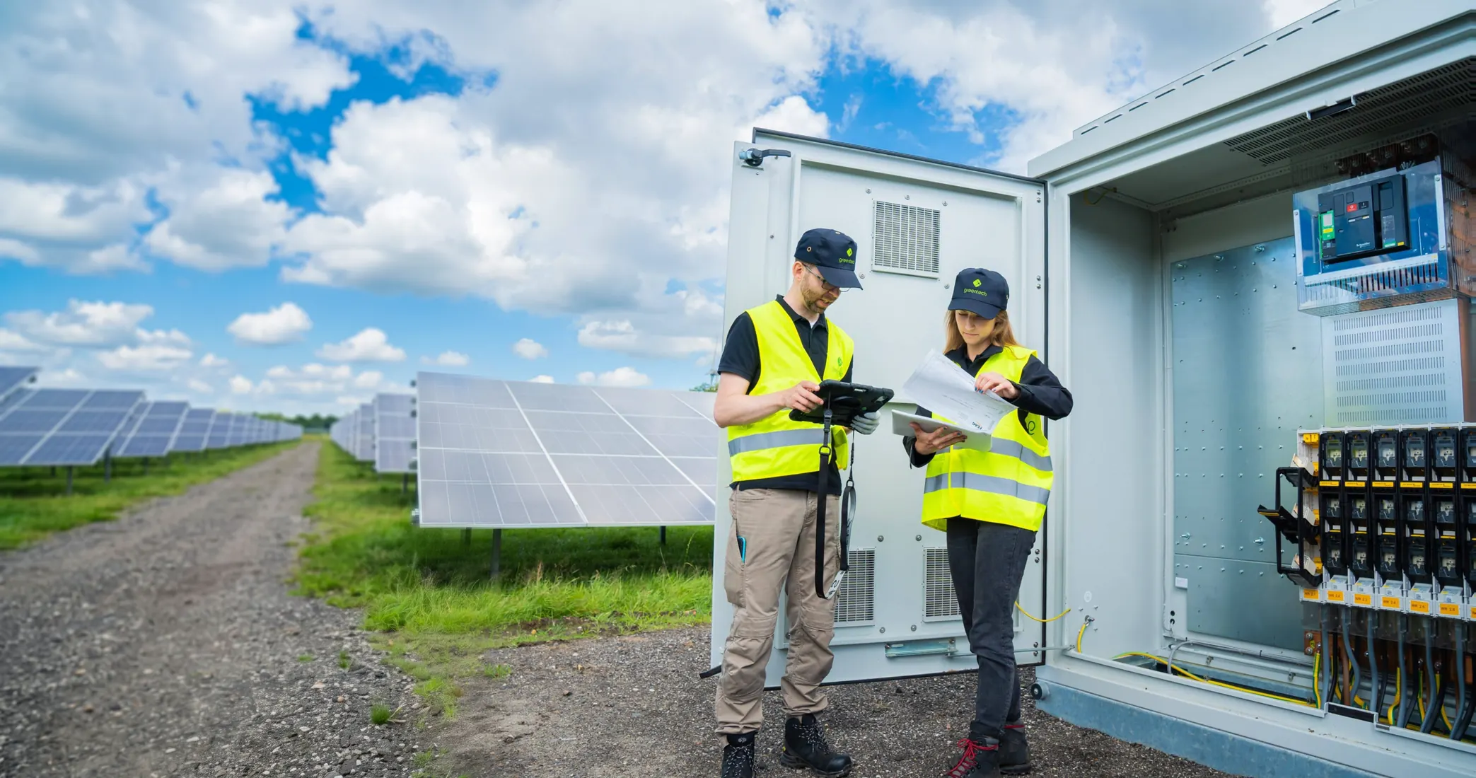 Two greentech technicians in high-visibility vests check a tablet and a test report in front of an open switchgear cabinet, with solar modules behind them.