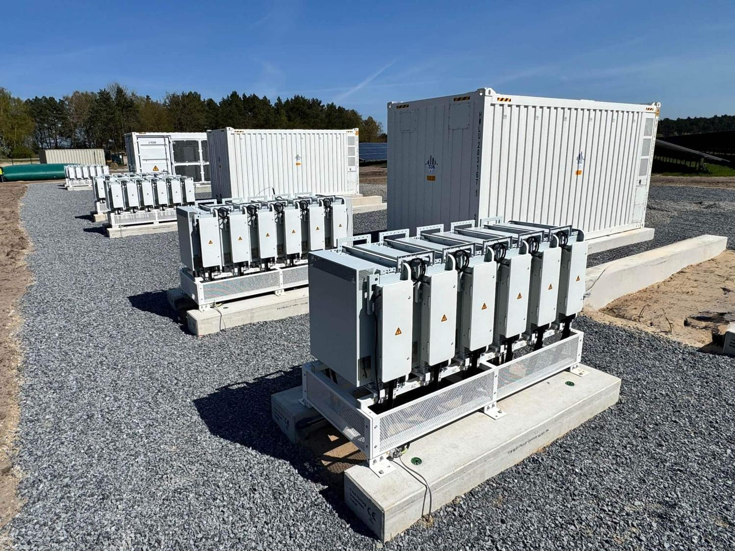 White battery storage containers and inverter units on a gravel bed under a blue sky, with solar modules behind.