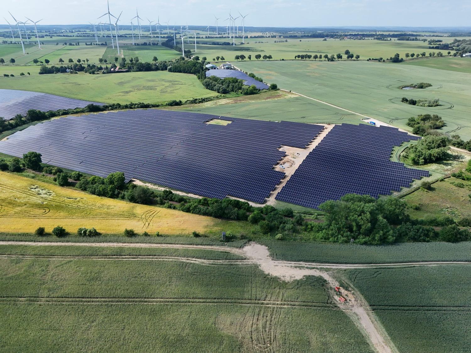 Aerial view of the Schulzendorf solar park between fields, with wind turbines on the horizon.