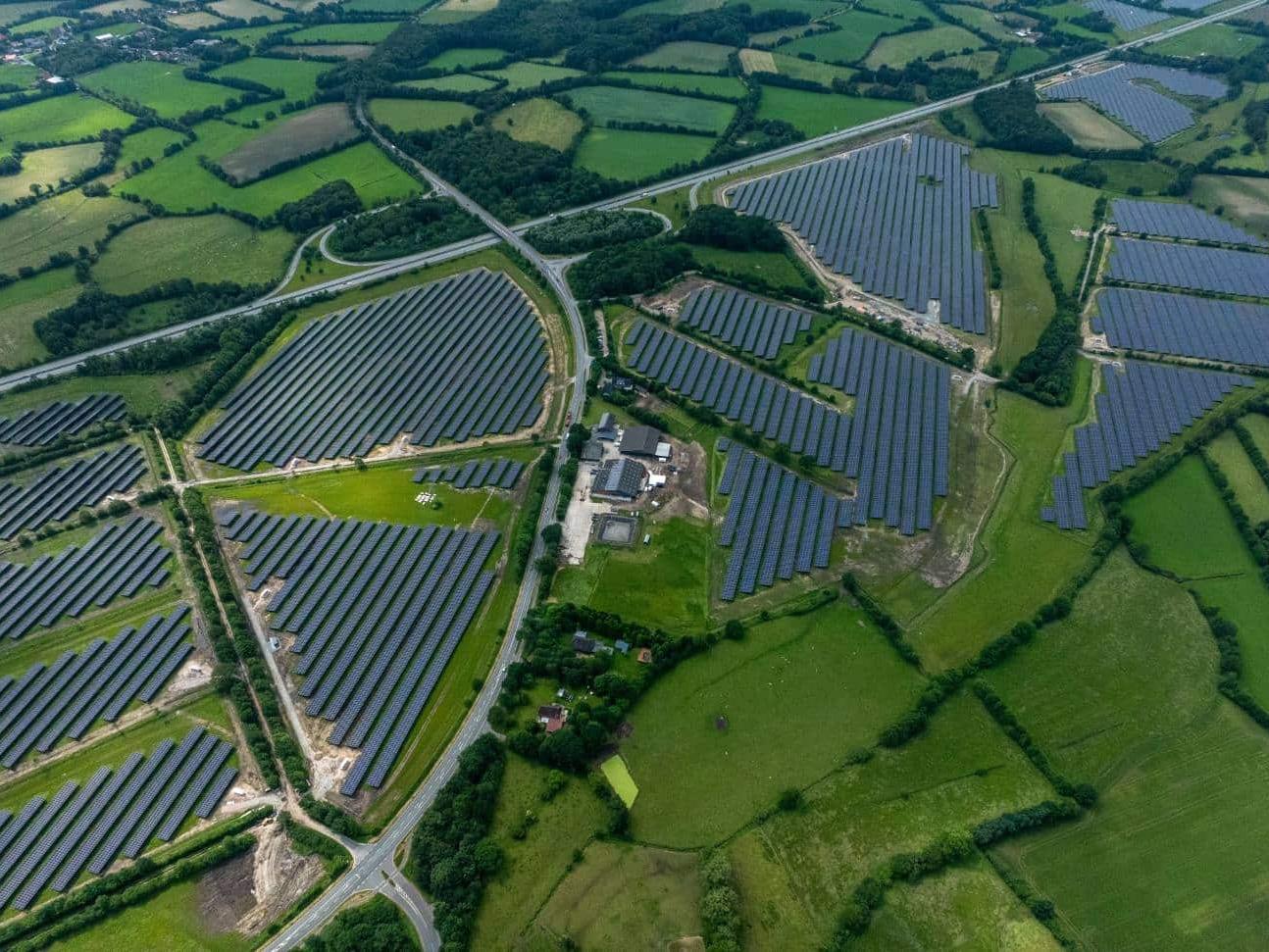 Aerial view of several solar fields between green pastures, hedgerows and a motorway junction.