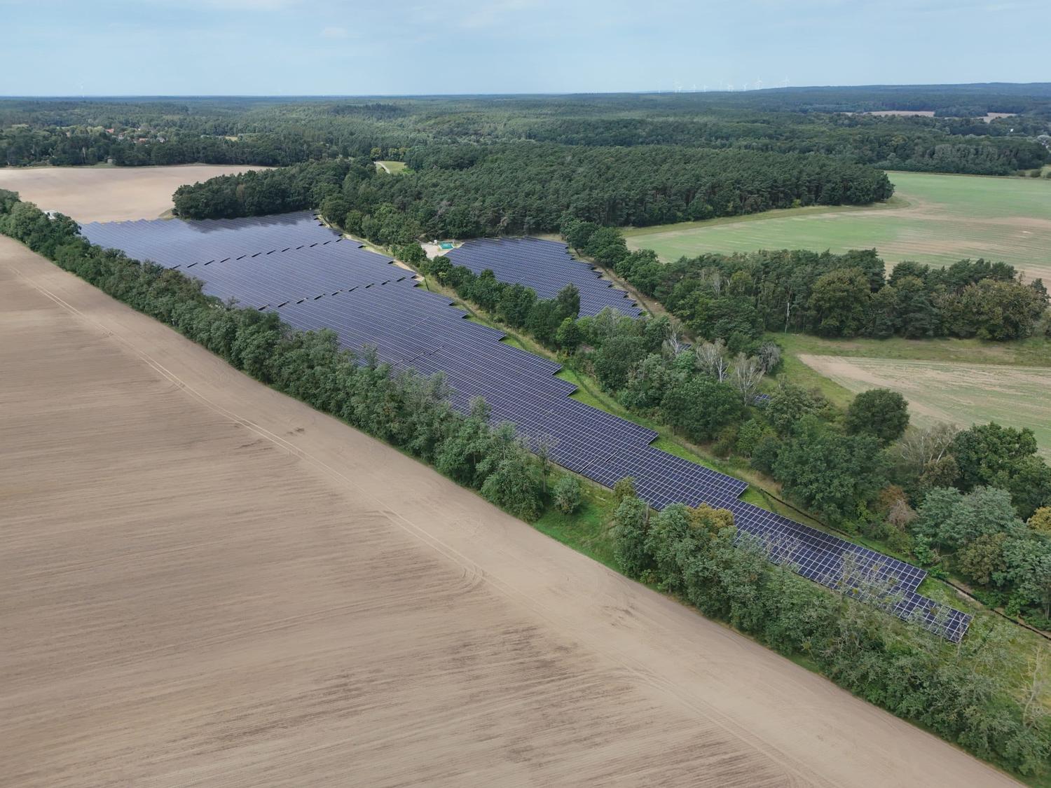 Aerial view of a solar park bordered by a row of trees, forest and a bare field.
