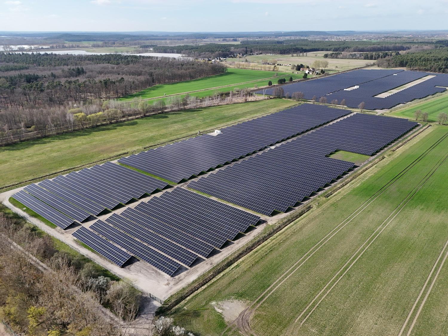 Aerial view of a large solar park in two blocks between green fields and woodland, with lakes on the horizon.