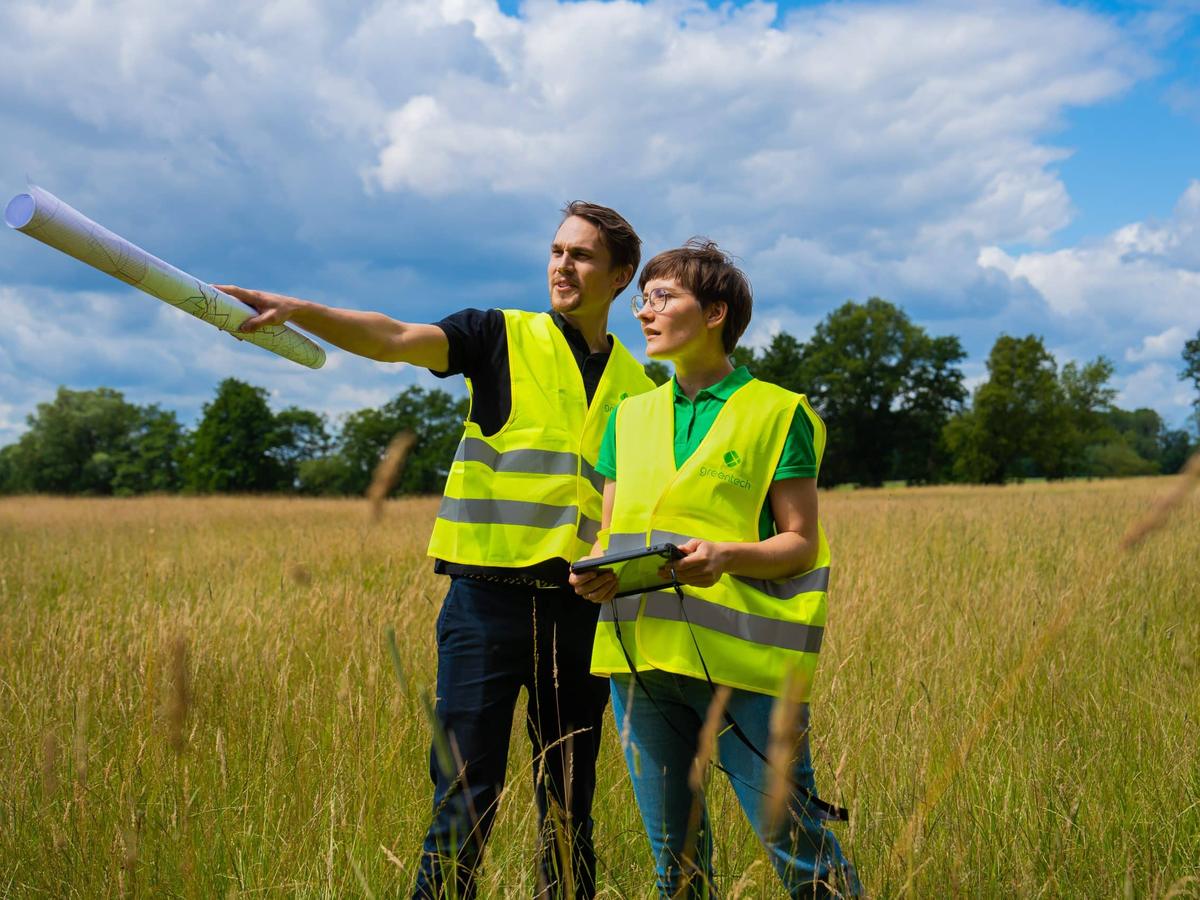 Two greentech employees in high-visibility vests on a meadow, one pointing across the site with a rolled plan